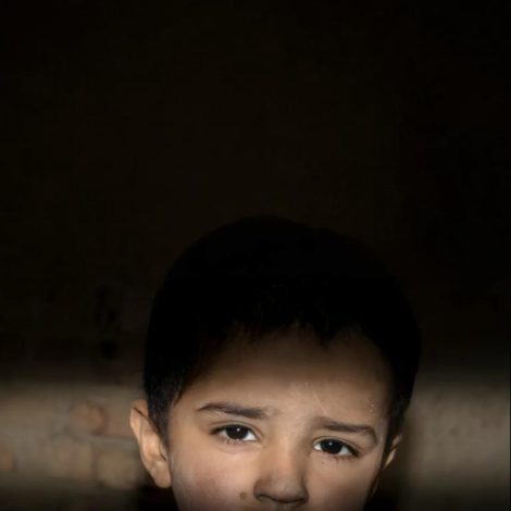 A Palestinian child's face partially illuminated by sunlight in a dark room, symbolizing hope amid war. Support Gaza orphans today. Help Palestinian Muslims Facing Genocide
